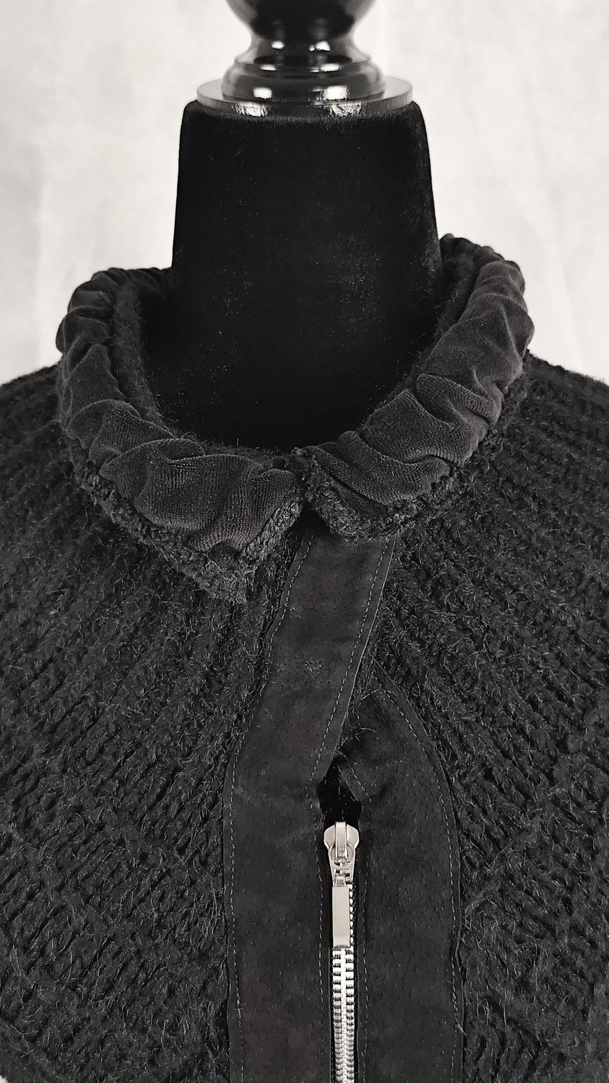 A close-up of a black textured knit cardigan displayed on a black mannequin torso, featuring a ruffled collar and a front zipper.