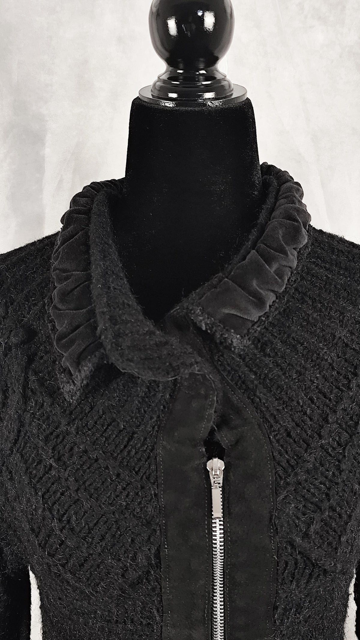 A black textured knit jacket with a velvet ruffled collar and a black zipper, displayed on a black mannequin torso against a light gray background.