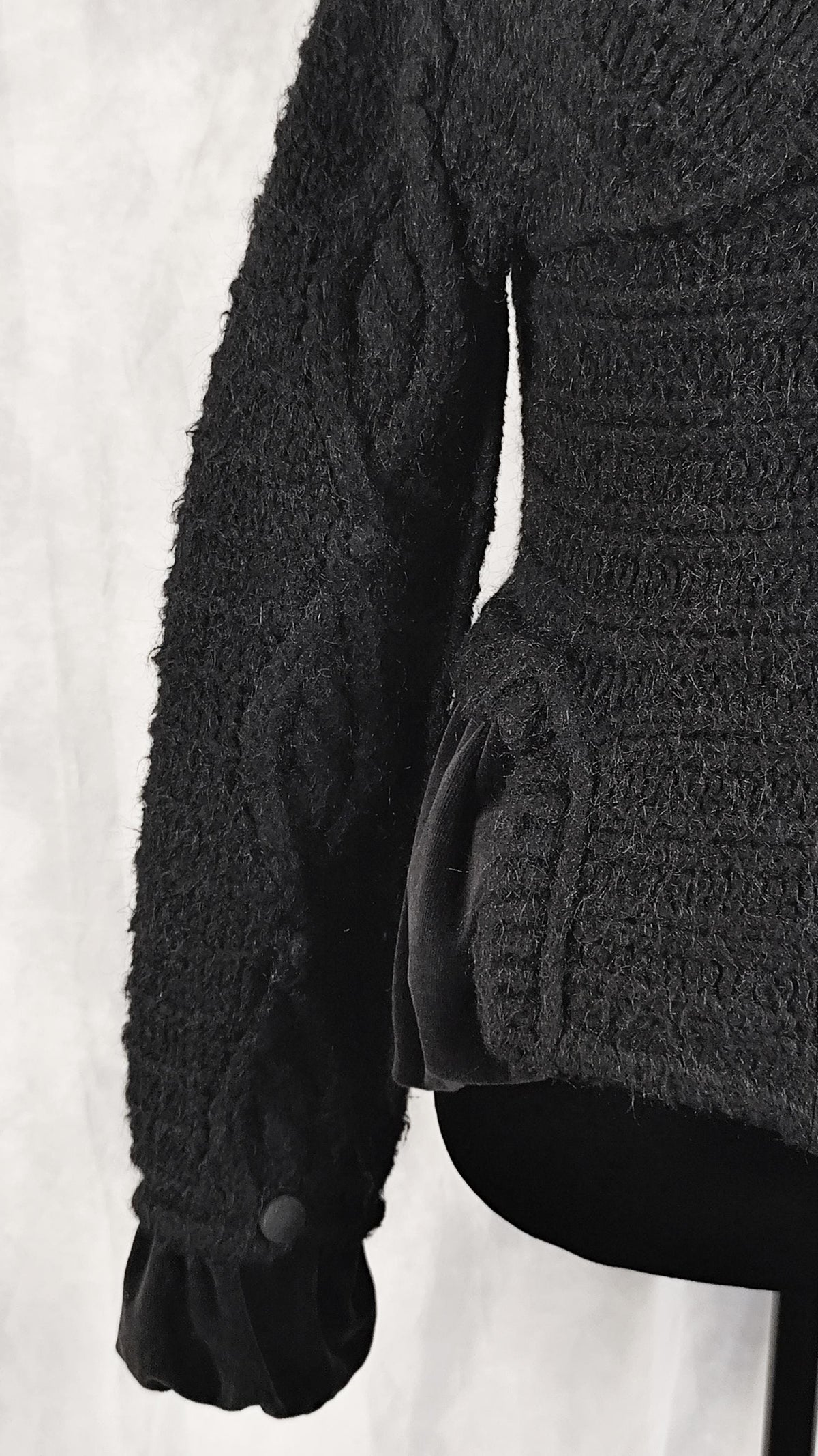 Close-up of a black textured knit sweater with a ribbed waist and a black velvet peplum hem, shown on a mannequin against a light gray background.