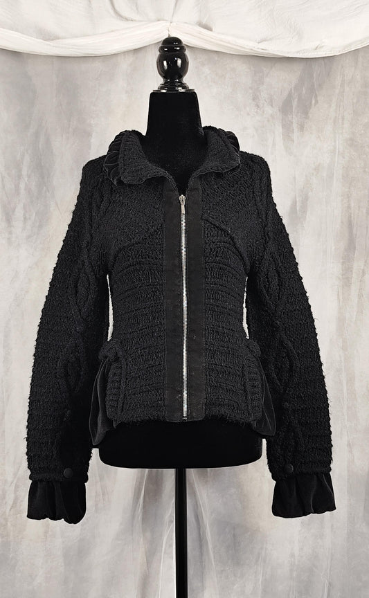Front view of a black textured zip-up knit jacket with a high collar, displayed on a black mannequin torso against a draped gray fabric backdrop.