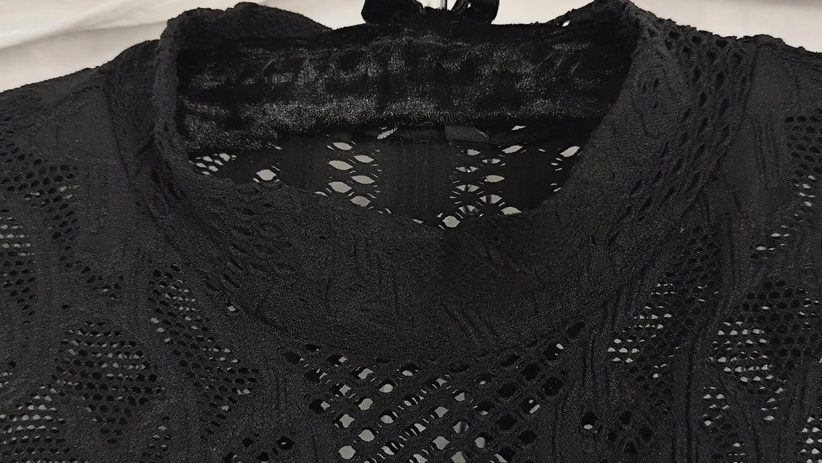 Close-up of a black lace dress with an openwork crochet pattern and a wide collar, shown against a light neutral background.