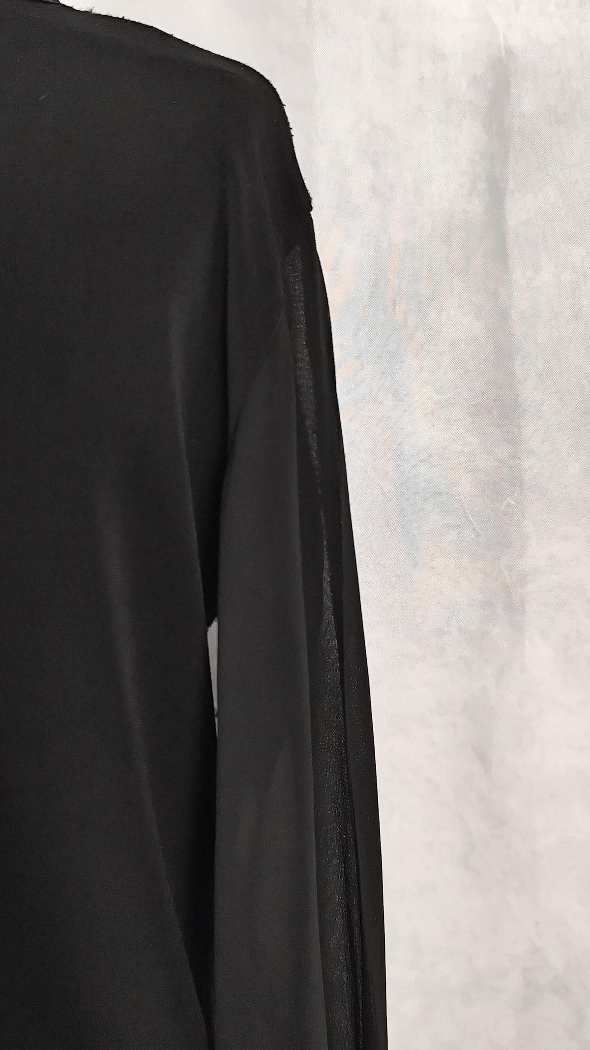 A close-up of a black long-sleeve cardigan with a sheer side panel, shown against a textured light gray backdrop.