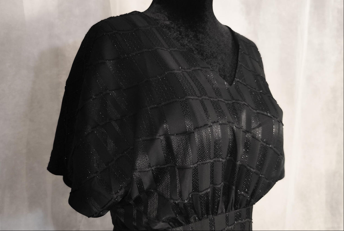 A black textured dress displayed on a black mannequin torso against a light gray fabric backdrop.