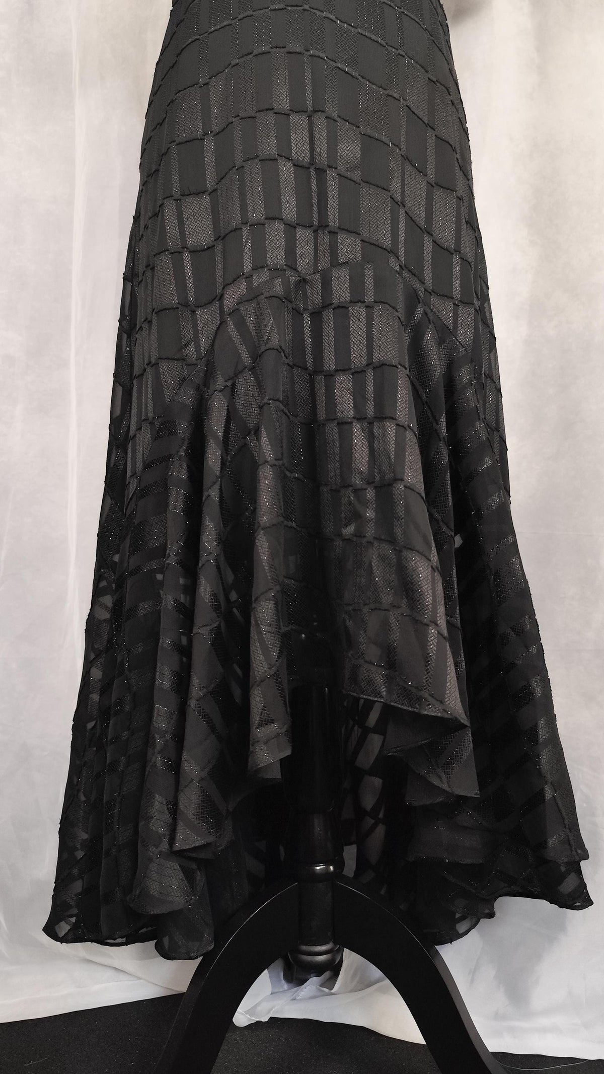 A black tiered ruffled skirt with a textured geometric pattern, displayed on a black dress form against a white fabric backdrop.