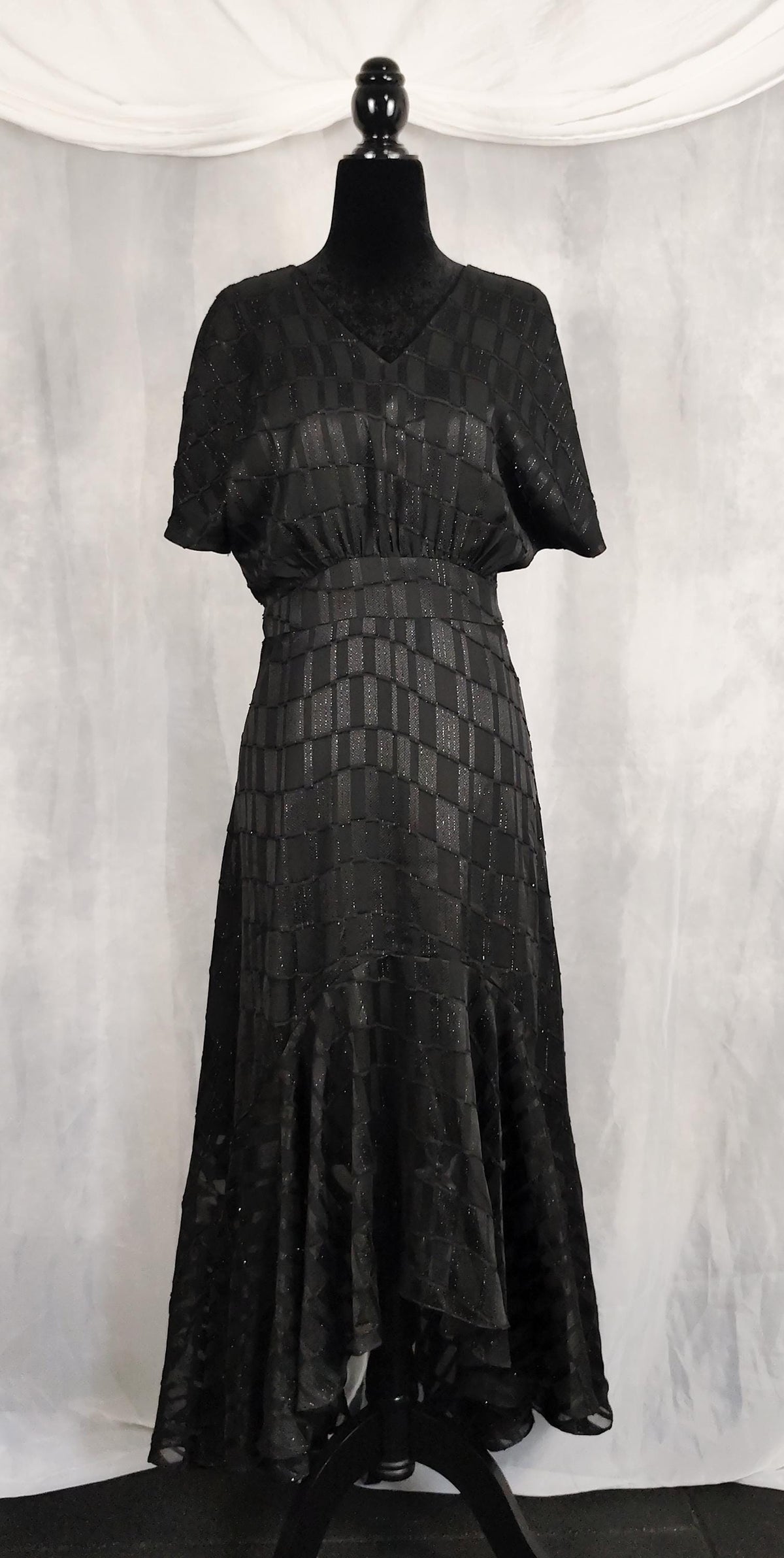 Front view of black mullet dress with short sleeves and a textured geometric pattern, displayed on a black mannequin against a draped white fabric backdrop.