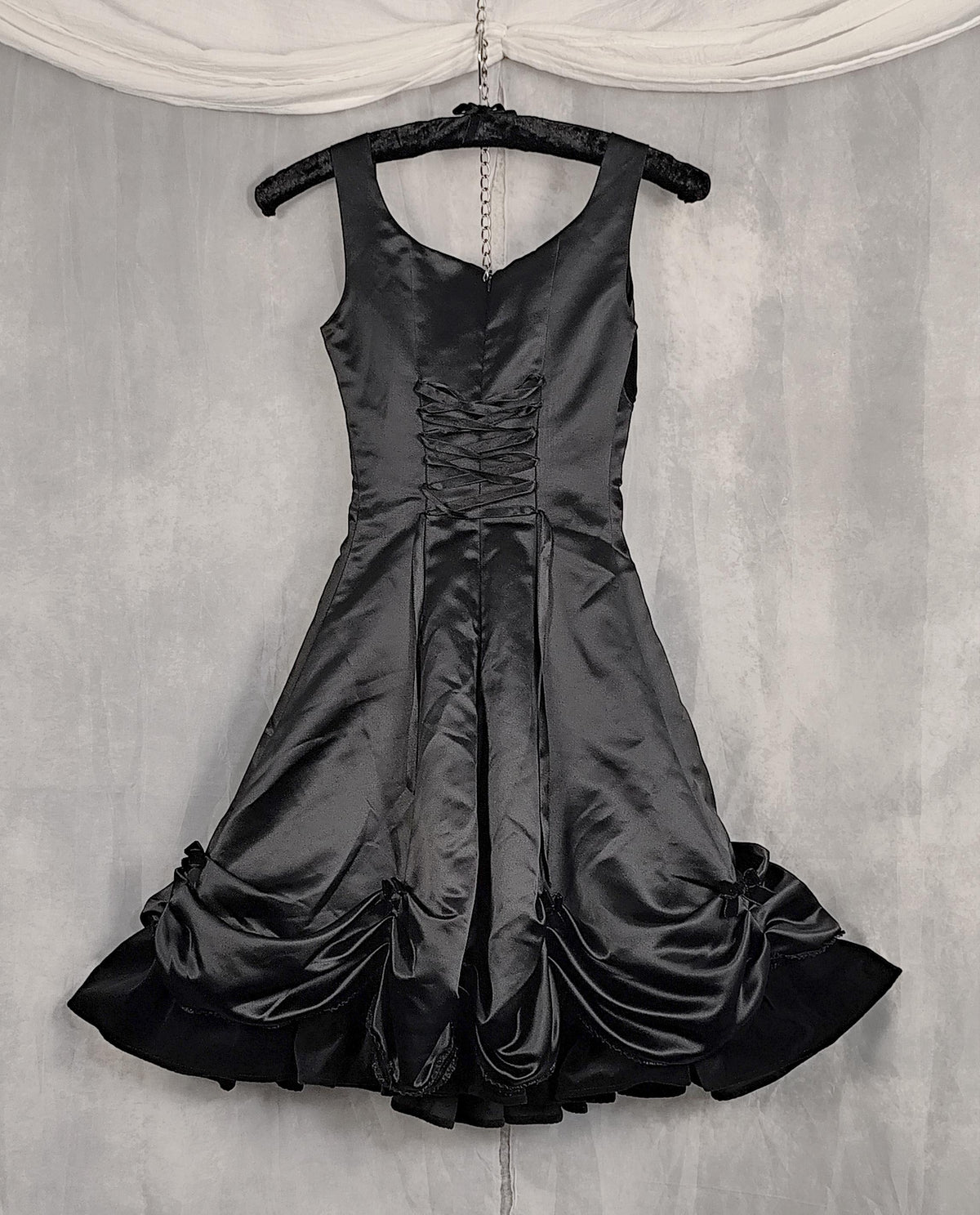 Back view of a black satin sleeveless dress with a lace-up corset bodice and gathered skirt, hanging on a black hanger against a textured gray backdrop with draped white fabric at the top.