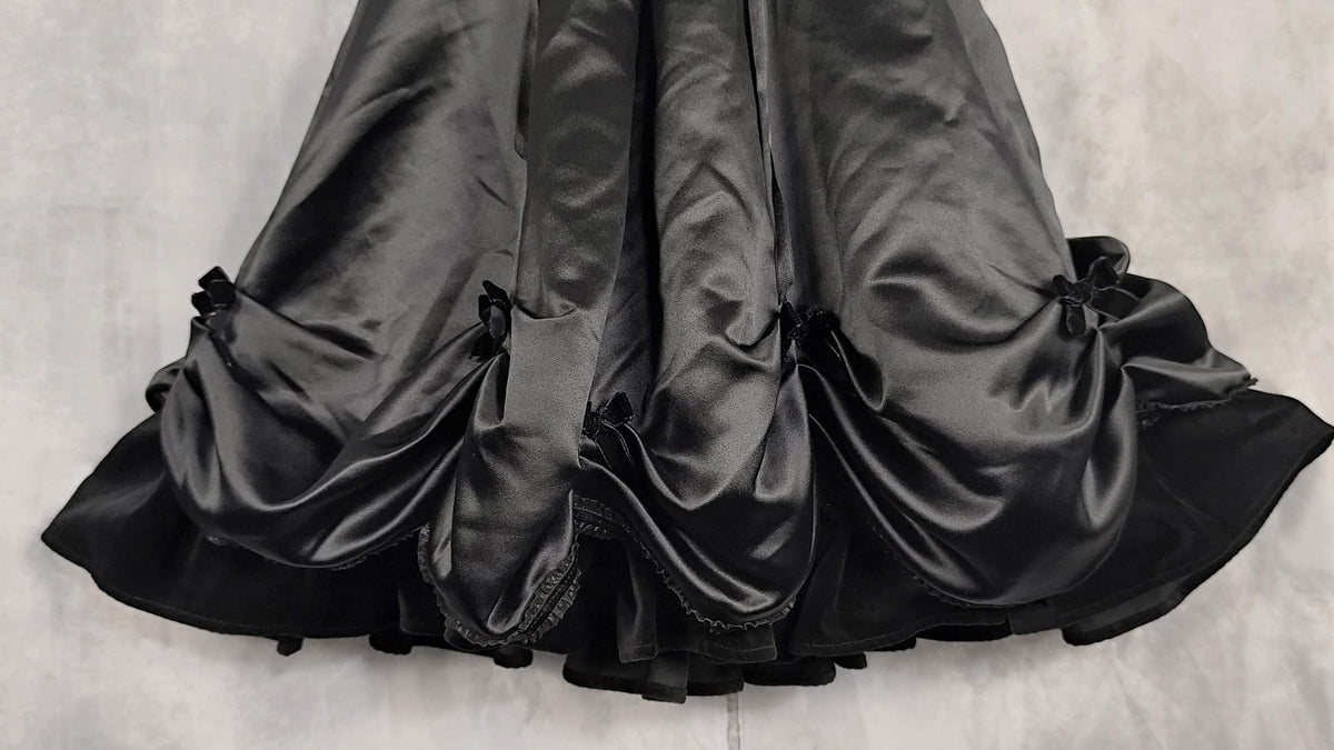Black satin ruffled skirt from a satin dress with gathered pleats and small velvet bow details, laid flat on a light gray textured background.
