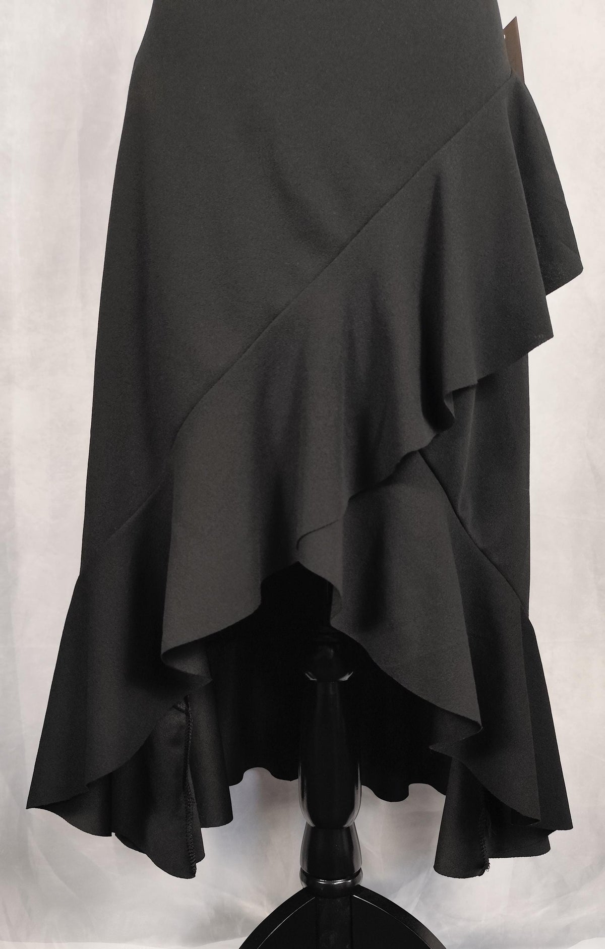Close-up of a black high-low ruffled skirt displayed on a black dress form against a white fabric backdrop.