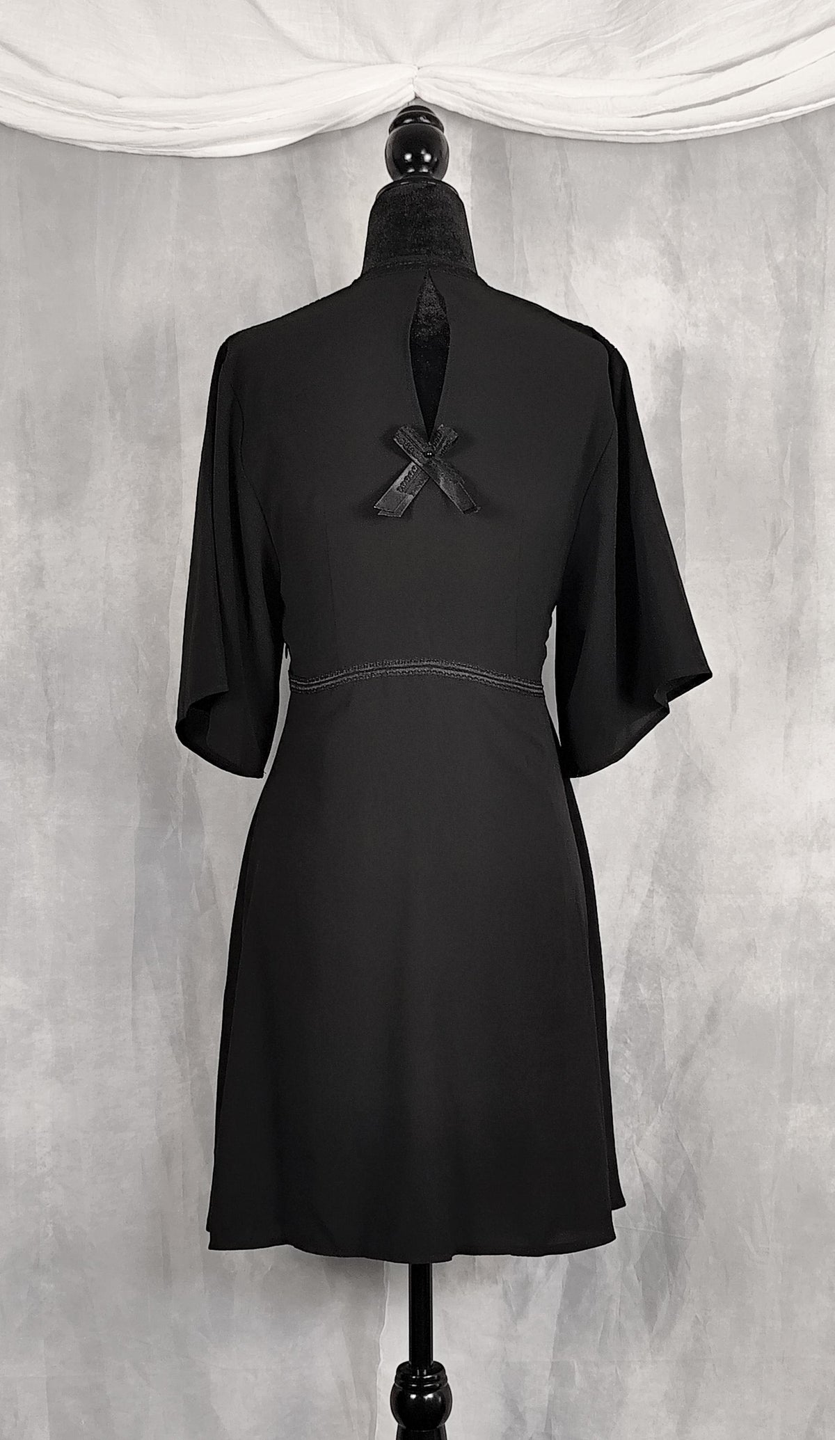 Back view of a black short-sleeved dress displayed on a black dress form mannequin, showing the back view with a keyhole cutout and bow detail, set against a gray textured backdrop with a draped white fabric at the top.