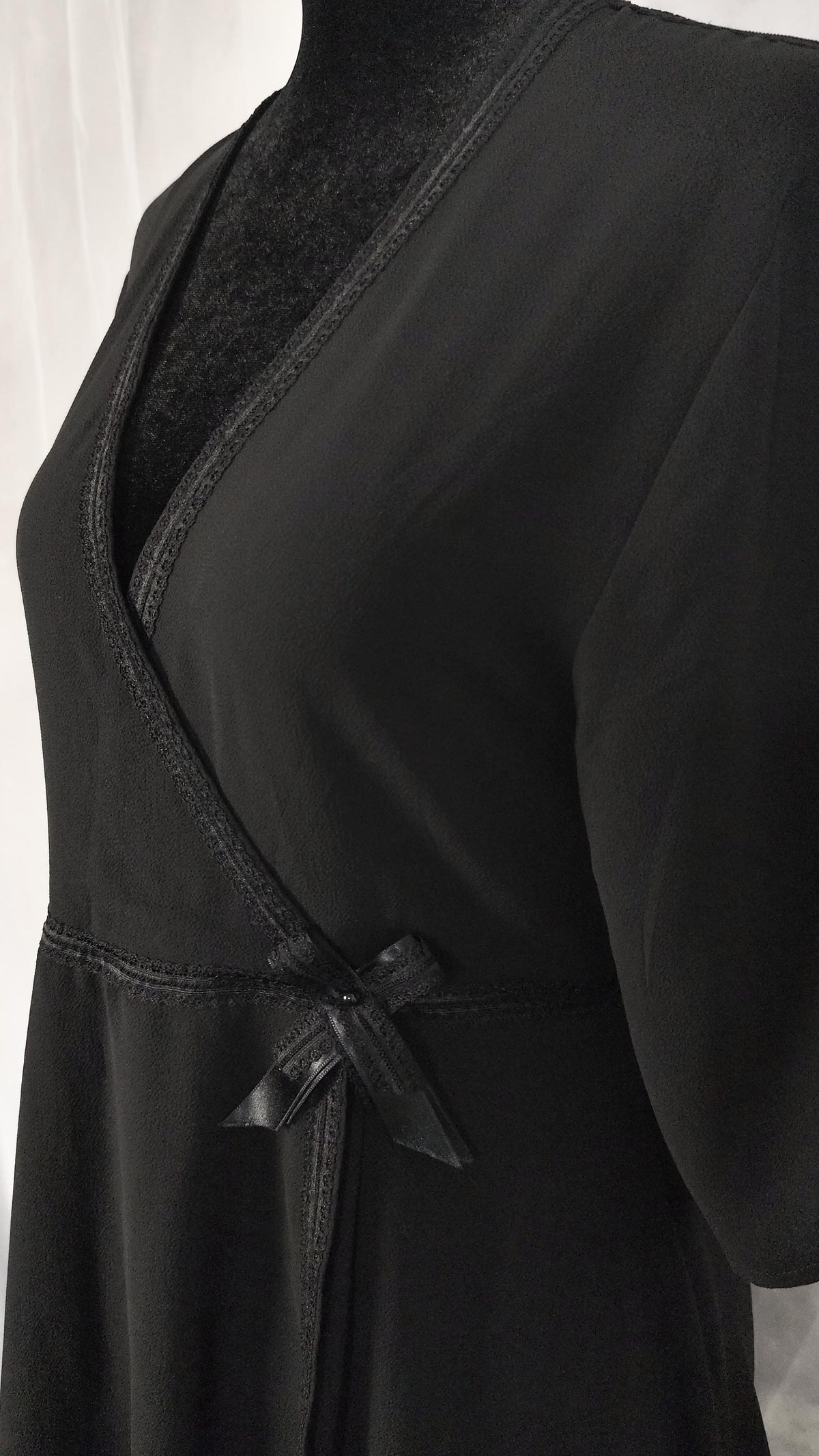 A close-up of a black wrap-style dress with a deep V-neck, short sleeves, and a small bow detail at the waist, shown on a mannequin against a light background.