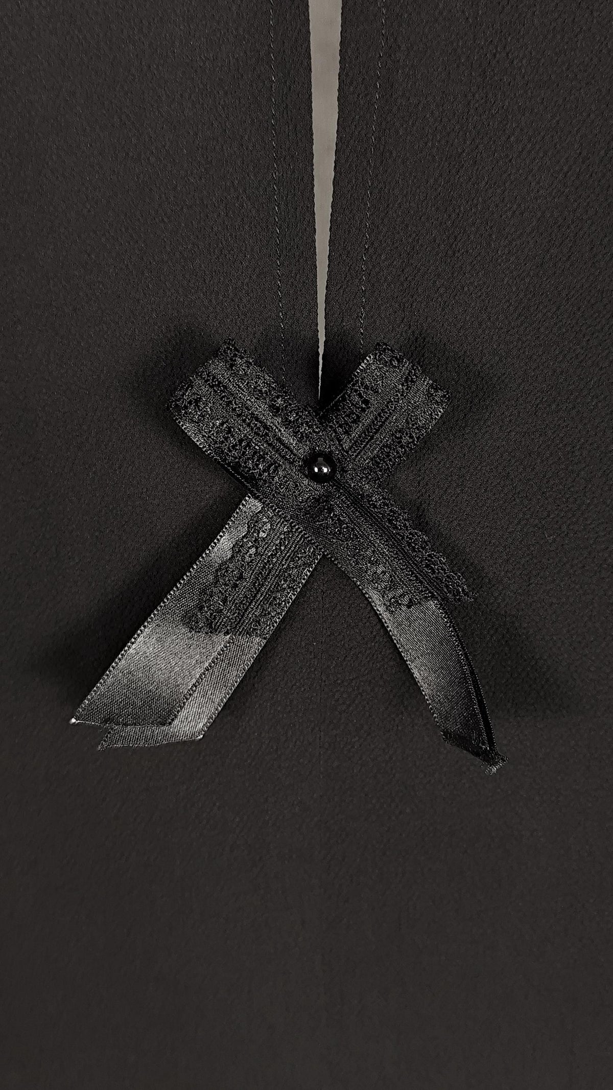 A close-up of a black dress featuring a decorative black lace ribbon bow with a small black bead at the center, set against a dark charcoal background.