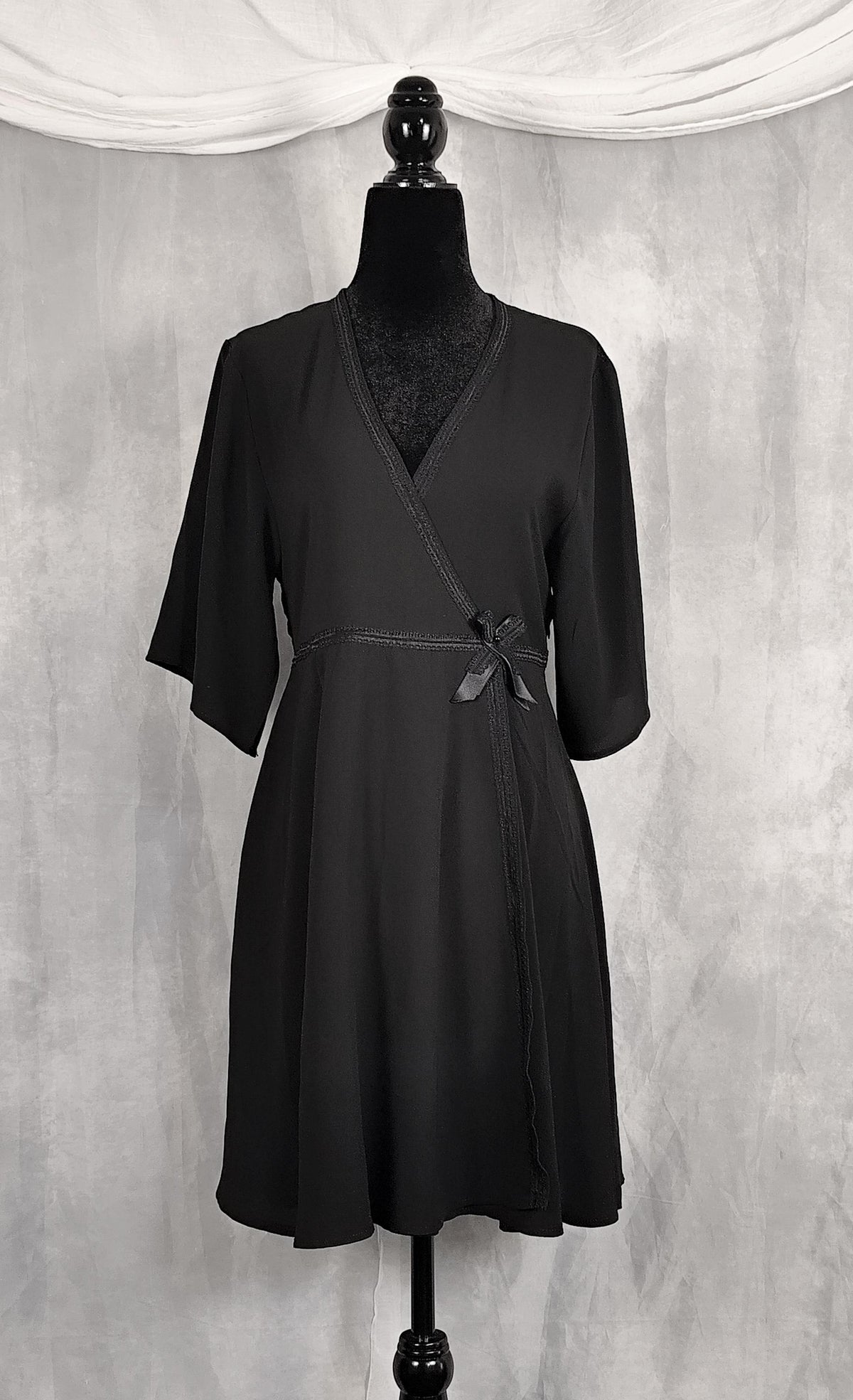 Front view of a black wrap-style dress displayed on a black mannequin against a textured gray backdrop with draped white fabric at the top.