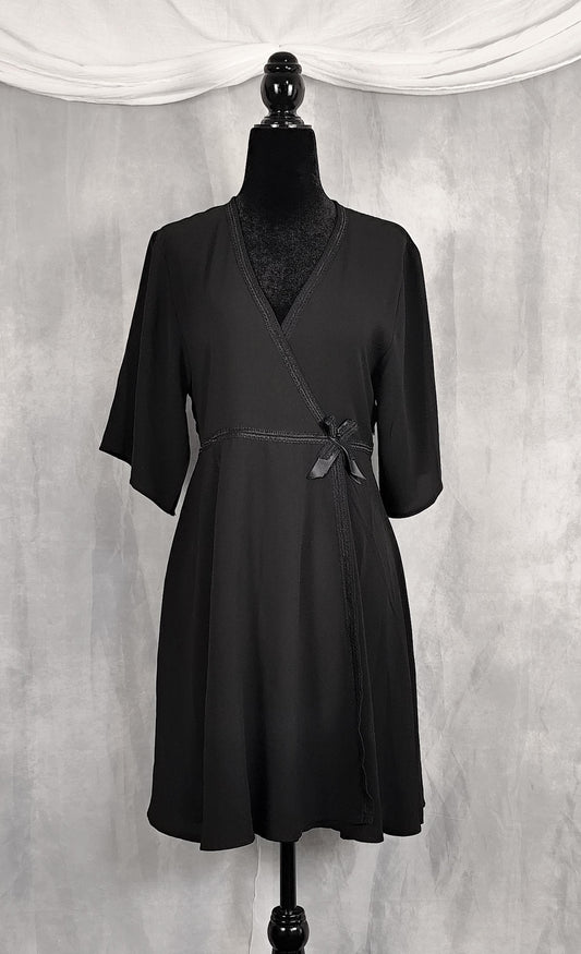 Front view of a black wrap-style dress displayed on a black mannequin against a textured gray backdrop with draped white fabric at the top.